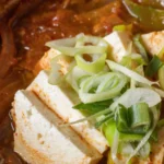 Kimchi Jjigae: O Que É e Como Fazer o Ensopado Coreano | Comidas Asiáticas: Ensopado coreano kimchi jjigae com pedaços de tofu e vegetais em panela de barro