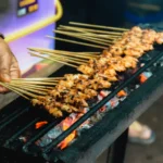 Satay: O Que É e Como Fazer Espetinhos com Molho de Amendoim | Comidas Asiáticas: Espetinhos de frango satay dourando sobre uma churrasqueira a carvão na Indonésia